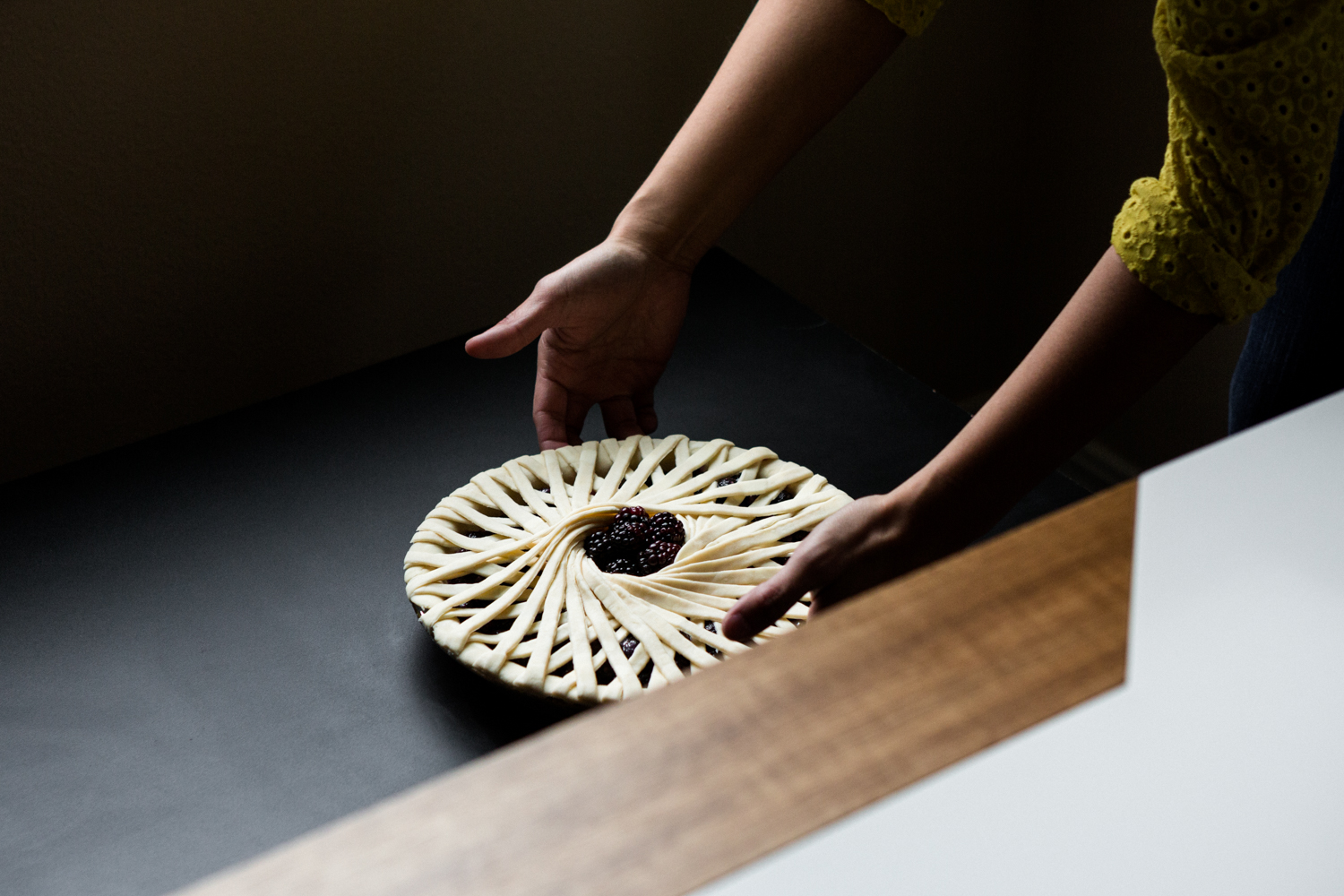 This image shows a person preparing a pie with a decorative lattice crust on a dark surface. The pie has a spiral pattern on the crust and contains dark filling, possibly berries. The person is wearing a yellow top and is using both hands to handle the pie. The background is dimly lit, and part of a wooden surface is visible on the right side of the image.