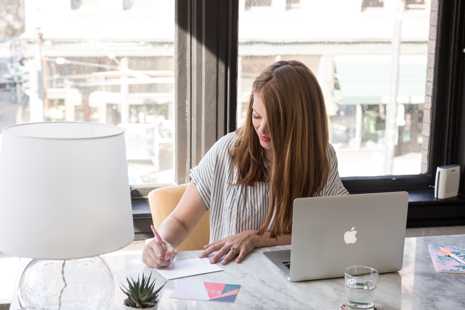 This image shows a person sitting at a desk in front of a large window, engaged in writing with a pen on a piece of paper. The desk is equipped with a lamp on the left side and an Apple laptop on the right side. The person is wearing a striped shirt and appears to be focused on their writing task. There is a small electronic device, possibly a speaker or a portable charger, placed on the window sill to the right. The background through the window shows an urban scene with buildings and some greenery.