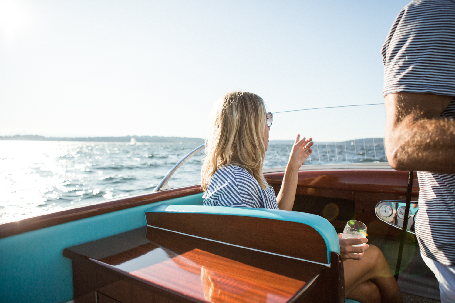 This image shows a person sitting on a boat, wearing a striped shirt and glasses, holding a glass, and gesturing with one hand. Another person is standing beside them, also wearing a striped shirt. The boat has a wooden and blue interior, with a polished wooden table in the foreground. The background features a clear blue sky and a body of water, suggesting a sunny day on the water.