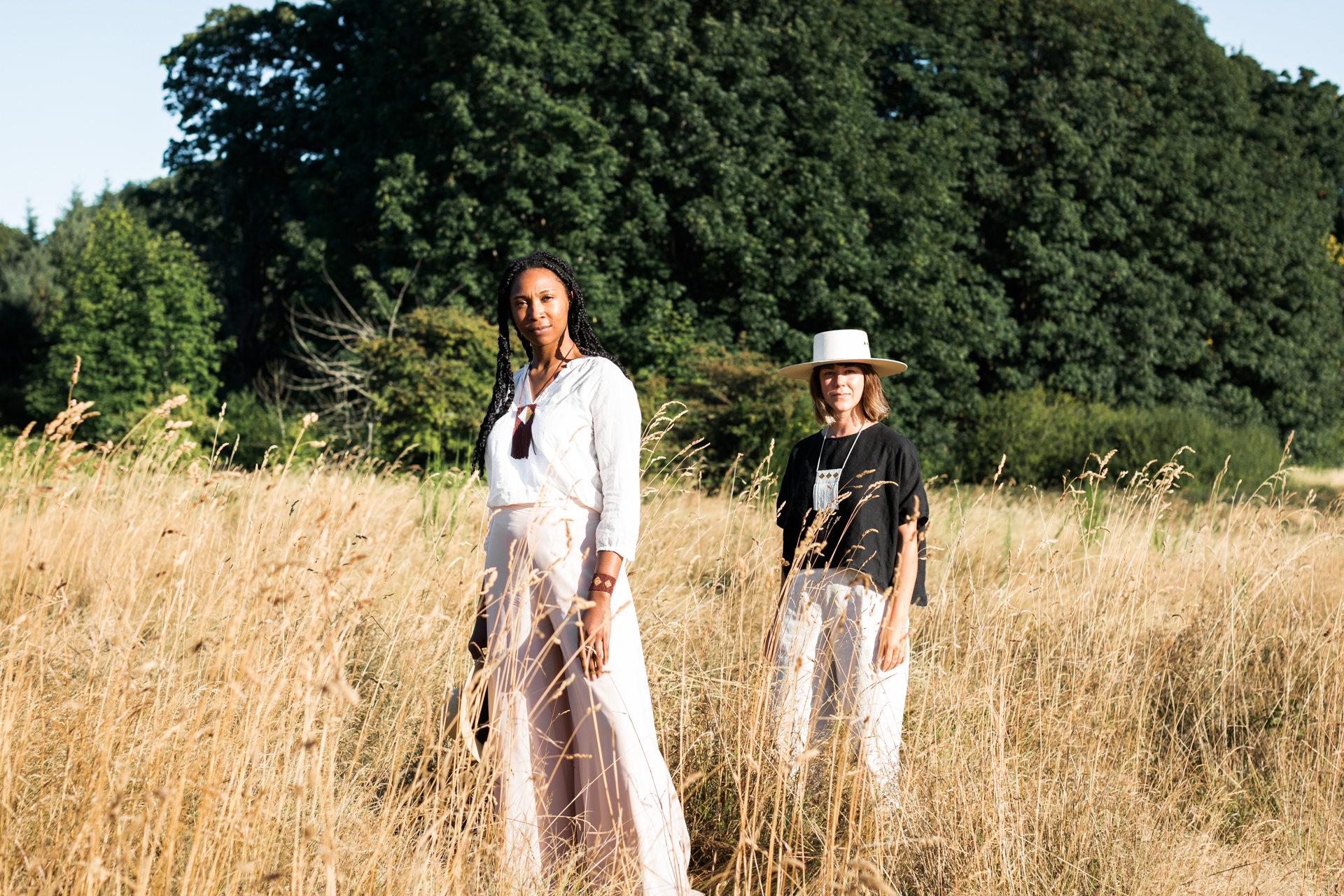 This image shows two individuals standing in a field of tall grass. The person on the left is wearing a white dress and a necklace, while the person on the right is wearing a black shirt, white pants, and a white hat. Both individuals appear to be posing for a photograph. The background features dense green foliage and trees, suggesting a natural, outdoor setting. The sky is clear and blue, indicating fair weather.