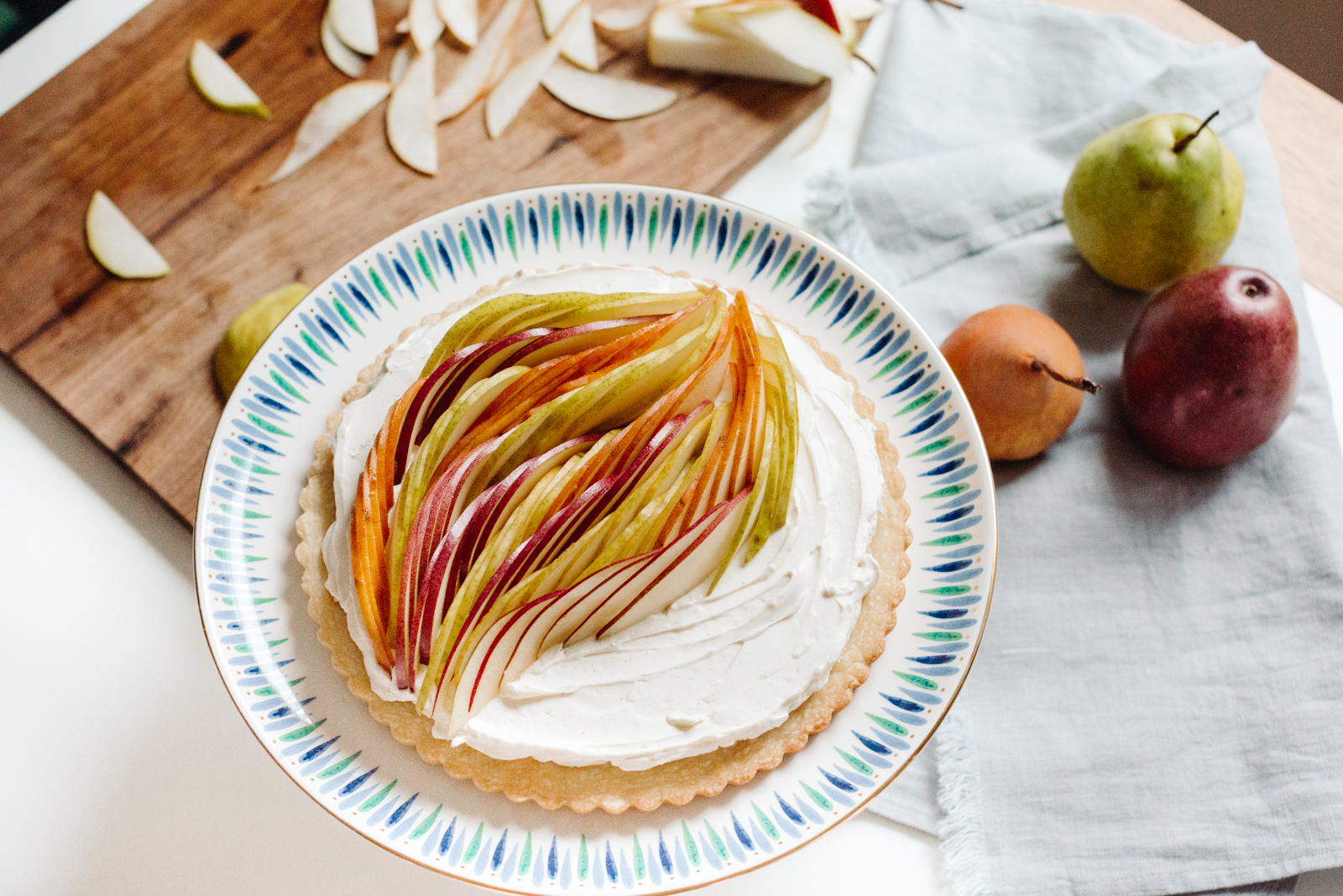 This image shows a tart with a white cream filling topped with sliced pears arranged in a decorative pattern. The tart is placed on a round plate with a colorful rim, featuring blue, green, and white stripes. The plate is set on a white surface, likely a table. Surrounding the tart are additional pears, some whole and some sliced, placed on a wooden cutting board and a light-colored cloth. The scene suggests a fresh and appetizing dessert presentation.