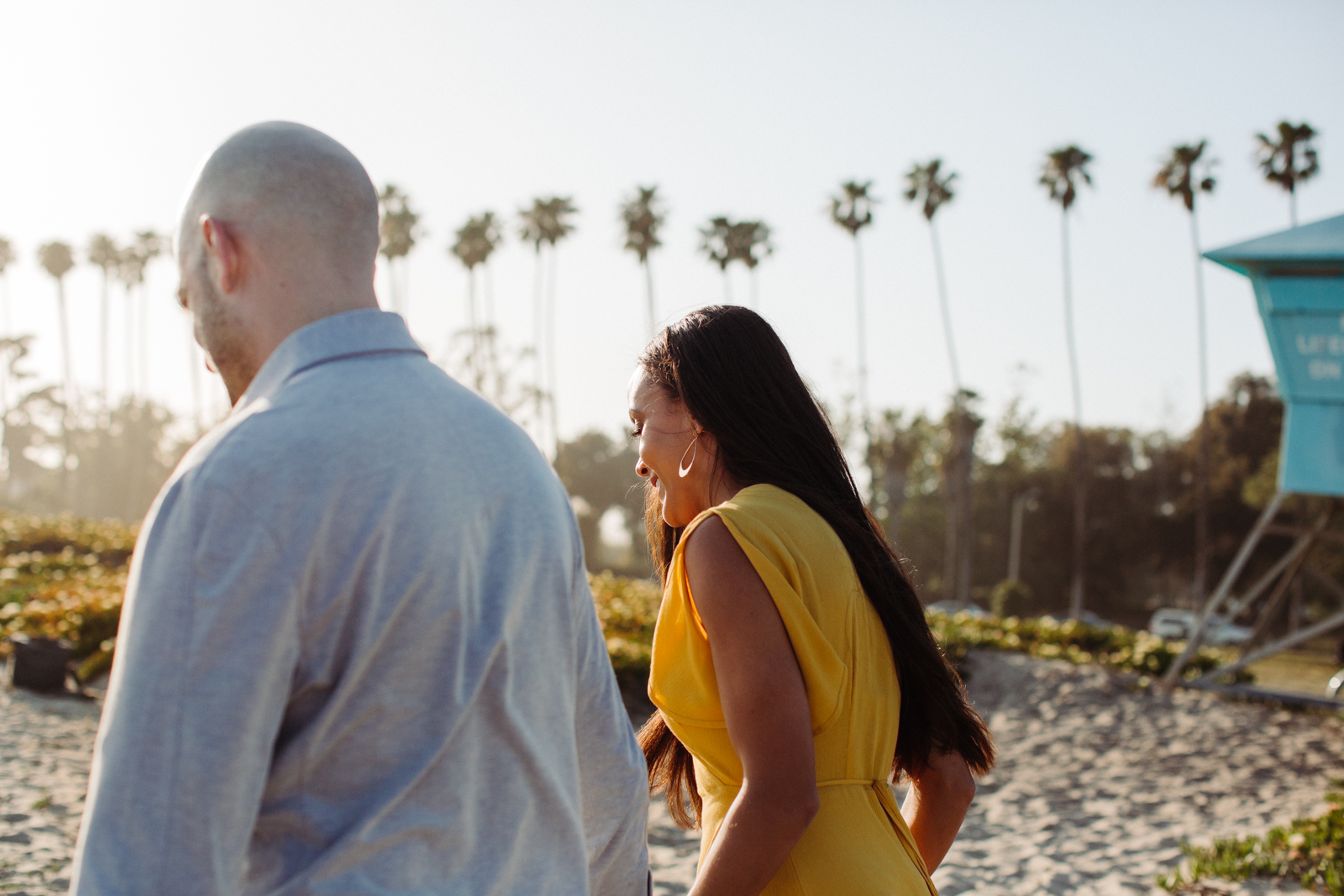 This image shows a couple walking along a sandy path, possibly at a beach. The man is wearing a light blue shirt and the woman is in a bright yellow dress. They appear to be engaged in a conversation, with the woman smiling. In the background, there are several tall palm trees, and a blue lifeguard tower is visible on the right side. The sky is clear and bright, suggesting it is daytime.