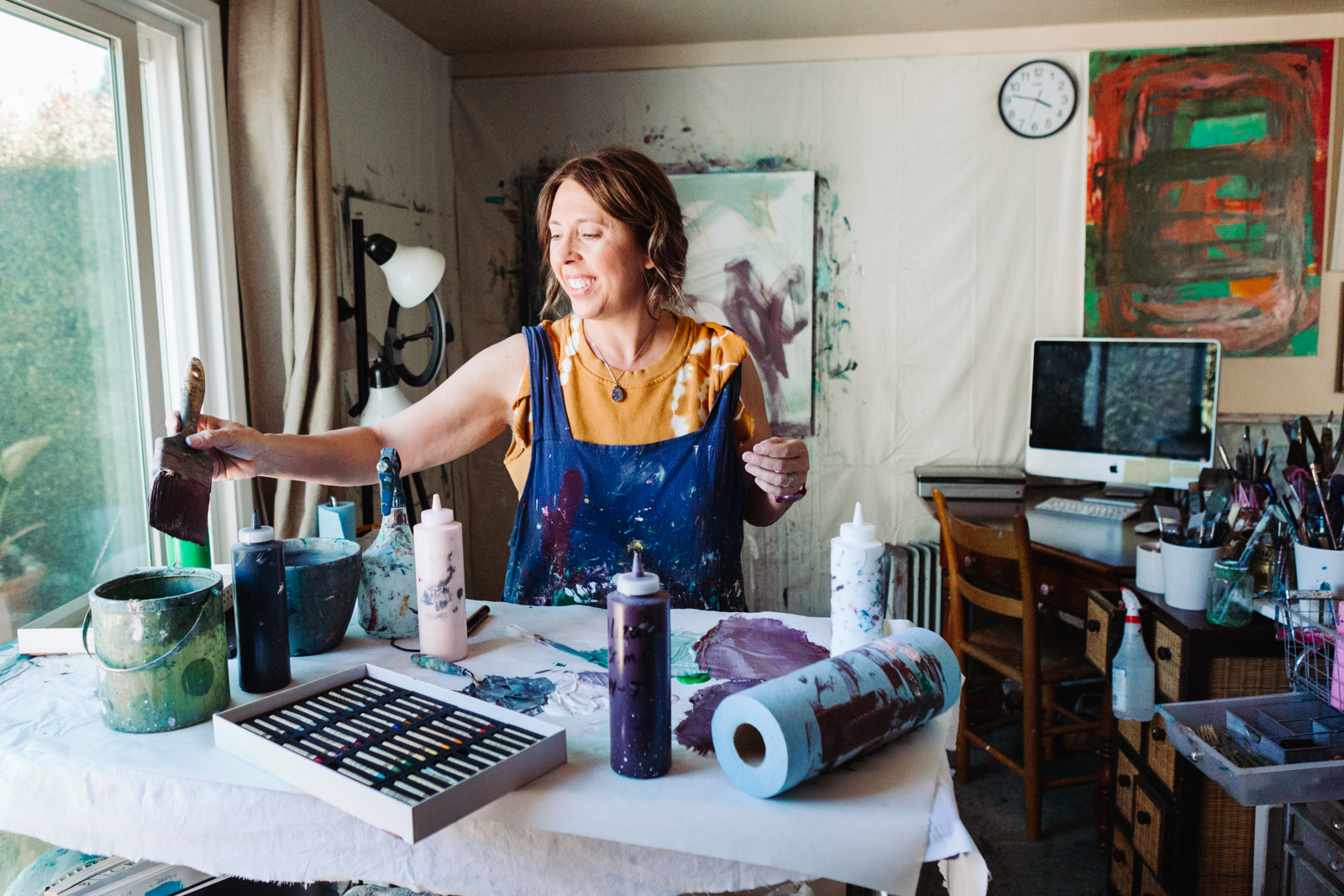 This image shows an artist working in a studio. The artist is standing behind a table covered with a white cloth, holding a paint palette and a brush. Various art supplies are scattered on the table, including paint bottles, a palette, and a roll of paper. In the background, there is a computer monitor on a desk, along with a keyboard and other art tools. A clock is mounted on the wall above the desk. The studio has natural light coming through a window on the left side.