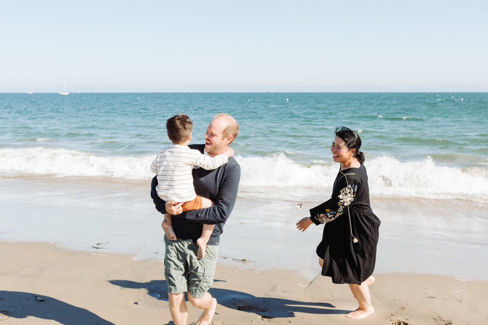 This image shows a family of three enjoying a day at the beach. A man is carrying a young child on his shoulders, both of them smiling. The child is wearing a striped shirt and shorts. A woman, dressed in a black dress with floral patterns, is walking towards them, also smiling. The beach is sandy with waves crashing in the background, and the sky is clear.