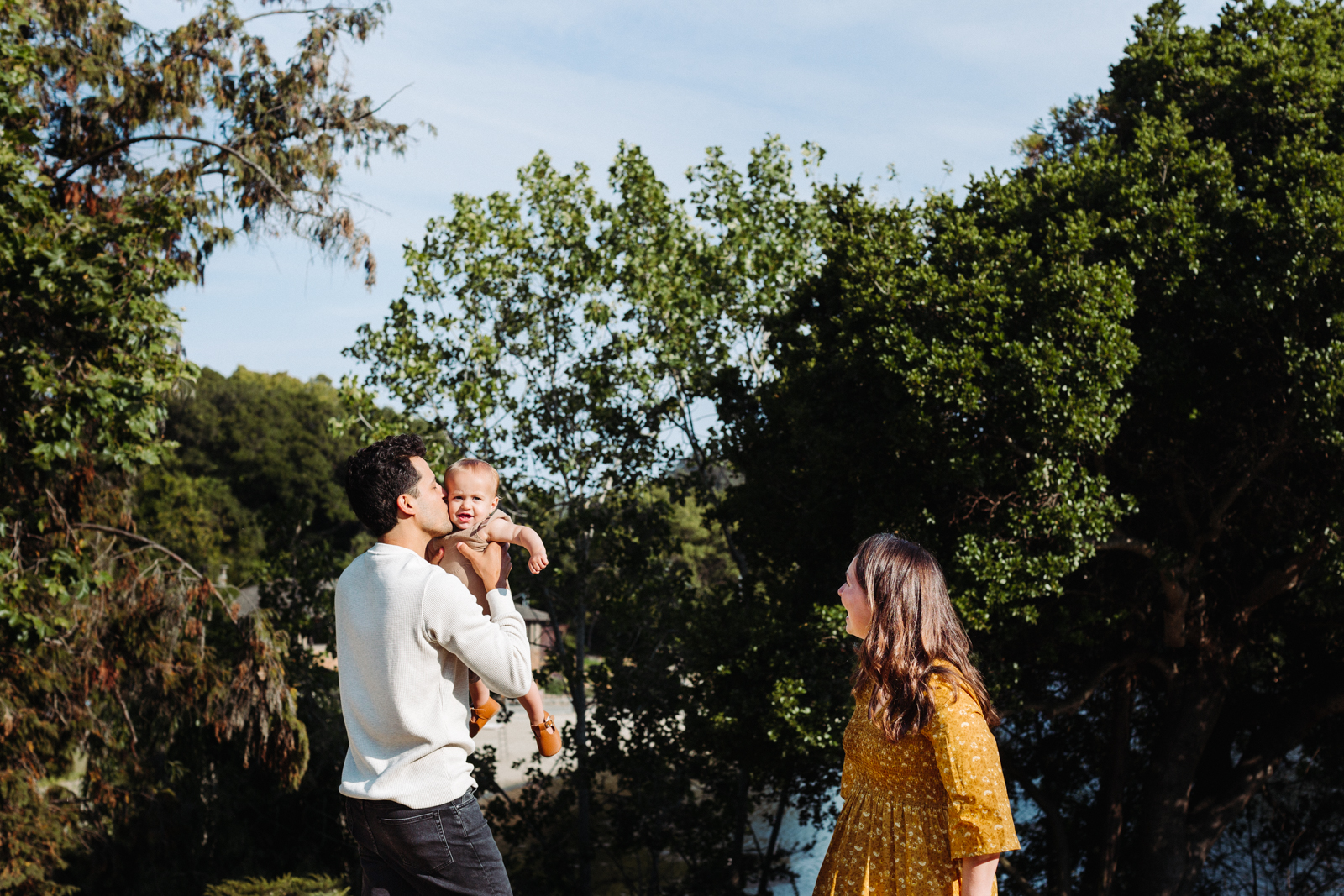This image shows a man holding a baby in his arms, with a woman standing nearby. The man is wearing a white long-sleeved shirt and dark pants, while the woman is dressed in a yellow dress. The baby is wearing a light-colored outfit. They are standing in an outdoor setting with lush green trees in the background, suggesting a park or a garden. The sky is clear and blue, indicating a sunny day.