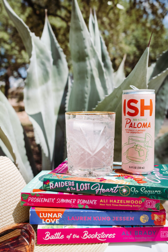 This image shows a stack of colorful books placed on a surface with a large agave plant in the background. The books have various titles and authors, including "Raiders of the Lost Heart" by Jo Segura, "Problematic Summer Romance" by Ali Hazelwood, "Lunar Love" by Lauren Kung Jessen, and "Battle of the Bookstores" by Ali Brady. In front of the books, there is a glass of a beverage with ice and a can of ISH Palomita non-alcoholic drink. The scene suggests a relaxed, leisurely setting with a focus on reading and enjoying a refreshing drink.
