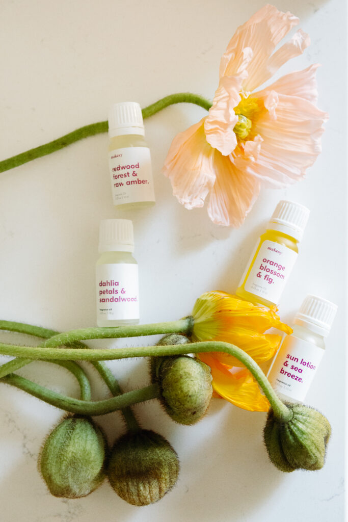 This image shows a collection of small bottles of scented oils arranged on a white surface. The bottles are labeled with various floral and natural scents such as "redwood forest & raw amber," "dahlia petals & sandalwood," "orange blossom & fig," and "sun lotion & sea breeze." Each bottle has a white cap and a label with the scent name and brand "makesy." The arrangement includes a few flowers and green stems, enhancing the natural theme of the display.