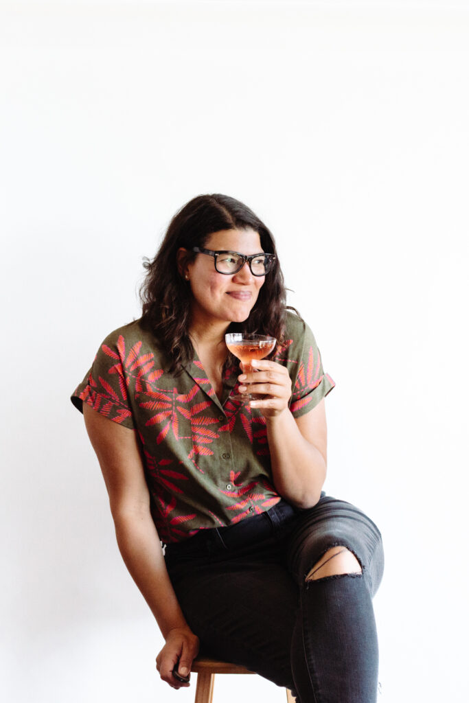 This image shows a person sitting on a stool against a plain white background. The person is wearing a green shirt with red leaf patterns and dark jeans with a visible tear on the right knee. They are holding a glass filled with a light-colored liquid, likely an alcoholic beverage, and are wearing glasses. The person appears to be posing for a photograph, with a slight smile on their face.