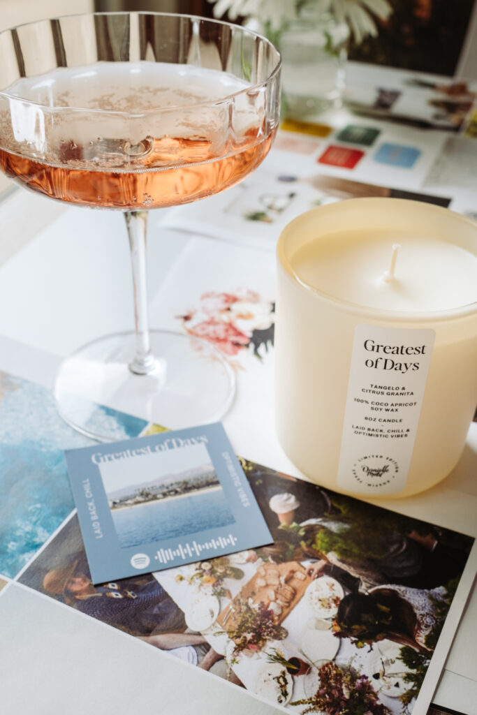 This image shows a table setting with various items. There is a glass of pink wine in a clear wine glass, a white candle with a label that reads "Greatest of Days" and mentions ingredients like tangelo, citrus granita, and coco apricot. Next to the candle is a blue card with the text "Greatest of Days" and a Spotify logo. There are also several photographs depicting people in outdoor settings, with one showing a picnic scene. The overall theme suggests a relaxed and enjoyable atmosphere, possibly for a special occasion or celebration.