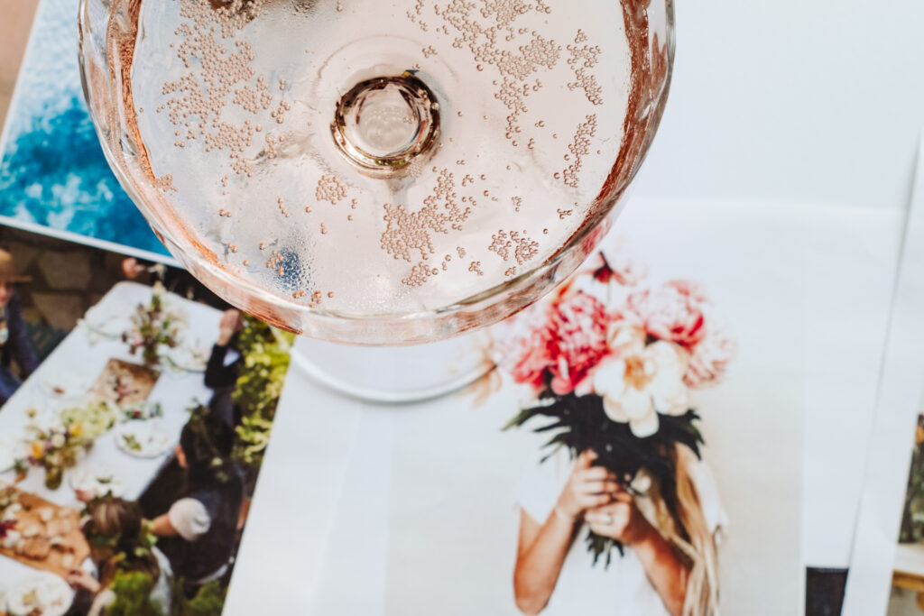 This image shows a close-up view of a glass filled with a sparkling beverage, possibly champagne or a similar drink. The glass is positioned over a collage of photographs, one of which features a person holding a bouquet of flowers. Another photograph depicts a dining table set with various dishes and flowers, suggesting a festive or celebratory occasion. The overall scene conveys a sense of elegance and celebration, with the sparkling drink adding to the festive atmosphere.