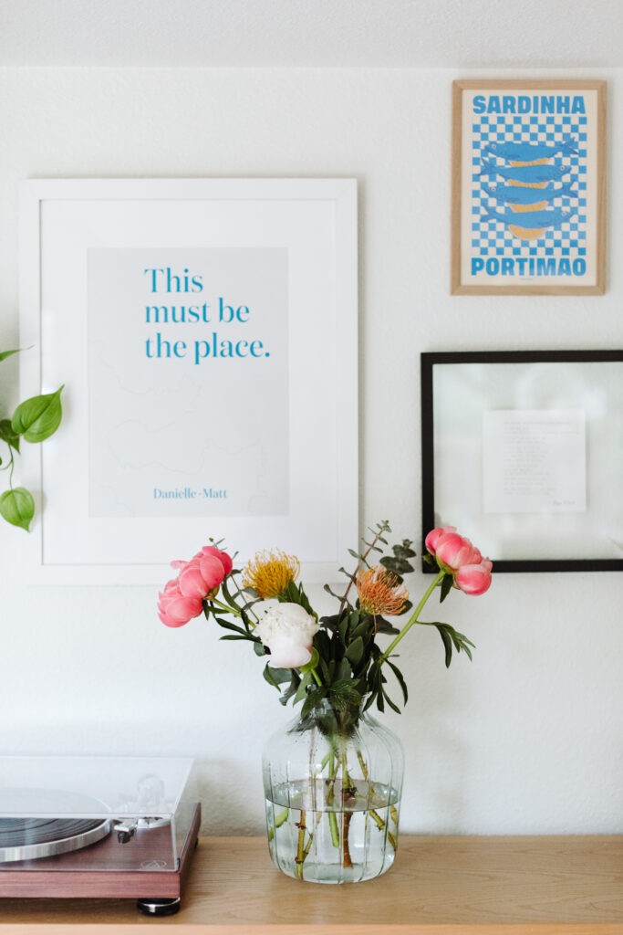This image shows a minimalist and aesthetically pleasing interior space. There is a wooden shelf against a white wall, holding a clear glass vase filled with water and a vibrant arrangement of pink and yellow flowers. Below the vase, a record player is partially visible on the left side of the shelf. Above the shelf, there are three framed items on the wall: a large framed print with the text "This must be the place." by Danielle Matt, a smaller framed poster with an image of sardines and the text "SARDENHA PORTIMAO," and a black-framed piece of paper with faint, illegible text. The overall scene conveys a calm and inviting atmosphere.