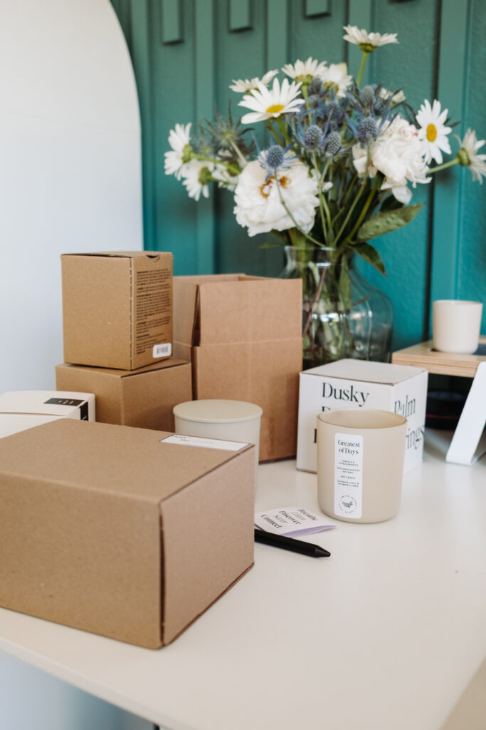 This image shows a neatly arranged table with various items. There are several brown shipping cardboard boxes of different sizes, stacked and placed on the table. A clear glass vase containing a vibrant arrangement of white flowers with green leaves is prominently displayed in the center. Next to the vase, there is a white cylindrical candle with the text "Greatest of Days" printed on it. Additionally, there is a white box labeled "Dusky" with some other text visible. A black pen and a piece of paper with printed text are also on the table.