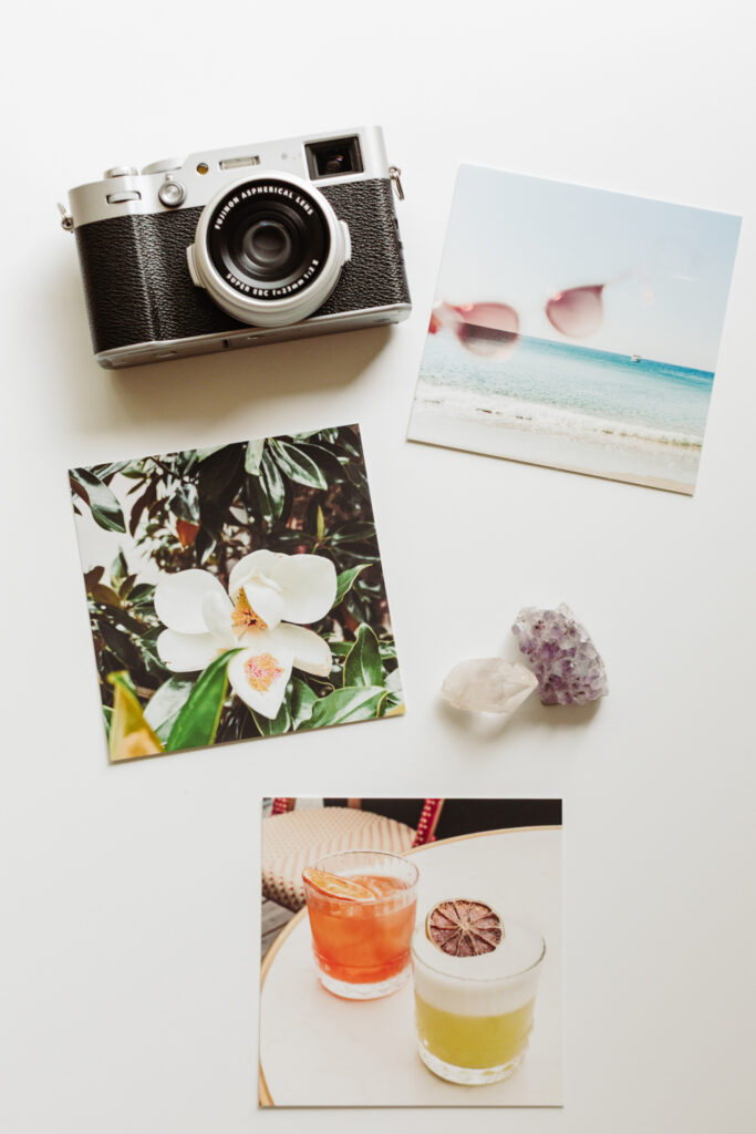 This image shows a collection of items on a white surface. There is a camera with a lens that reads "ASPHERICAL LENS" and "SUPER EBC 1:2.3/35mm." Next to it are three photographs. The first photograph depicts a beach scene with sunglasses superimposed. The second photograph shows a large white flower with green leaves. The third photograph features two drinks with garnishes on a table. Additionally, there are two crystals placed near the photographs.