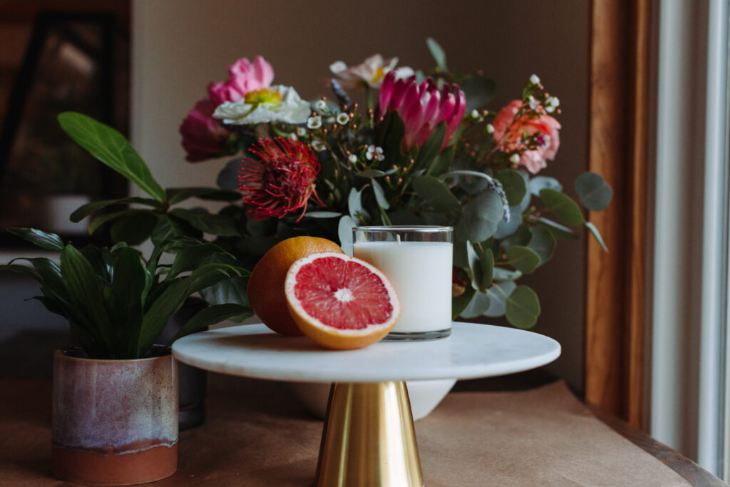 This image shows a decorative table setting with a white marble cake stand holding a sliced grapefruit and a glass of milk. Behind the cake stand, there is a large floral arrangement featuring a variety of colorful flowers, including pink, red, and white blooms, along with green foliage. To the left of the cake stand, there is a potted plant with broad green leaves. The table is covered with a light brown cloth, and the setting appears to be in a room with natural light coming through a window on the right side.
