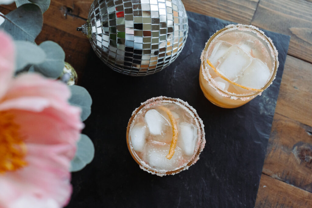 This image shows two glasses filled with a cocktail beverage, each topped with ice cubes and garnished with an orange twist. The glasses are placed on a dark rectangular surface, likely a table, which is set on a wooden surface. To the left of the glasses, there is a decorative silver disco ball and a bouquet of pink flowers with green leaves. The setting appears to be a casual and festive one, possibly for a gathering or celebration.