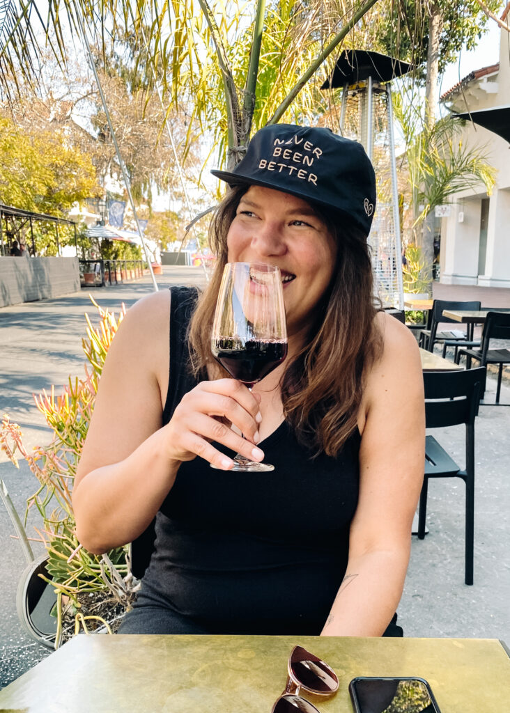 This image shows a person seated outdoors, holding a glass of red wine and smiling. The person is wearing a black cap with the text "NEVER BEEN BETTER" and a black sleeveless top. The setting appears to be a patio or outdoor dining area with tables and chairs. There are plants and trees in the background, and a smartphone is visible on the table in front of the person. The atmosphere suggests a relaxed and casual environment.