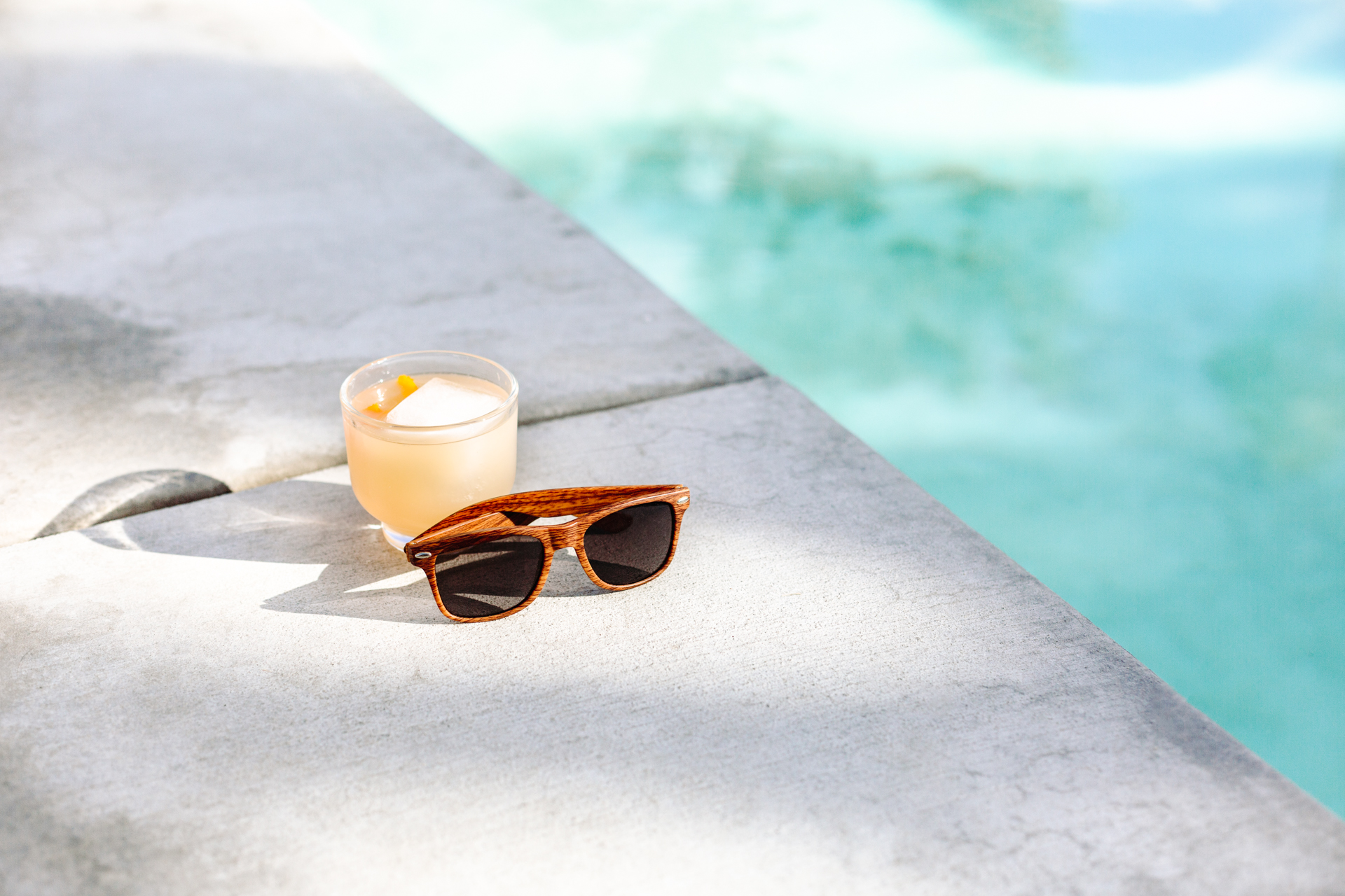 This image shows a pair of wooden sunglasses placed on a concrete surface next to a glass of a light-colored beverage with ice. The beverage appears to be a cocktail or a refreshing summer drink. The background features a swimming pool with clear blue water, suggesting a relaxing poolside setting. The sunlight casts shadows on the concrete, indicating it is a sunny day. The overall scene conveys a sense of leisure and relaxation.