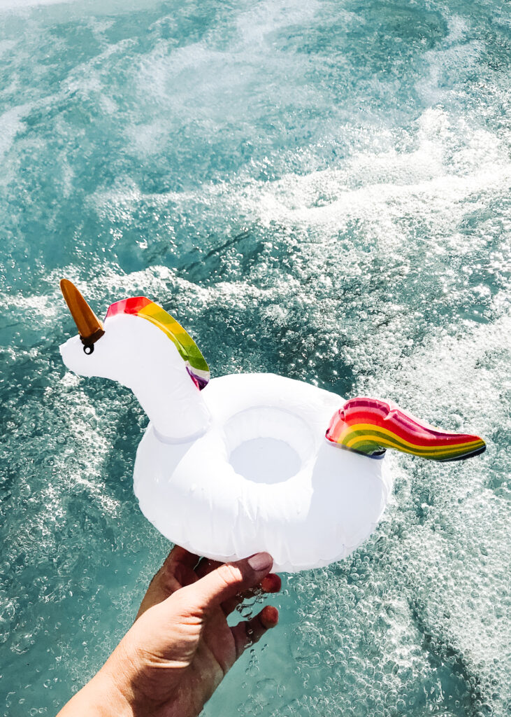This image shows a hand holding a white inflatable unicorn float toy in the water. The unicorn has a golden horn and a multicolored rainbow mane and tail. The water appears to be a pool or ocean with visible waves and bubbles. The hand holding the float is partially visible, showing fingers and part of the palm. The water is clear and blue, with light reflections indicating sunlight.