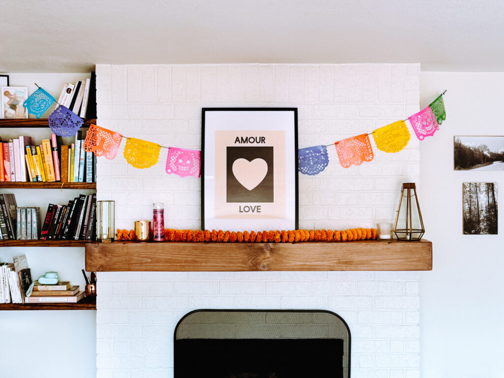 This image shows a cozy living room with a white brick fireplace. Above the fireplace, there is a framed poster with the words "AMOUR" and "LOVE" and a heart symbol. Colorful paper garlands are hung across the top of the fireplace. To the left, there is a bookshelf filled with various books, some of which have titles like "Collective Wisdom" and "THE LILIAD." On the right side, there are two framed pictures hanging on the wall. A decorative candle and a small vase are placed on the wooden mantel.