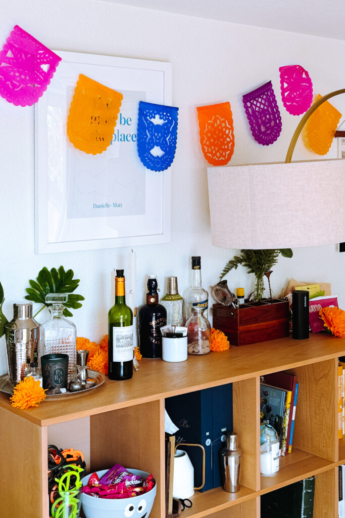 This image shows a room with a wooden shelf against a white wall. The shelf is adorned with various items including bottles, a lamp with a beige shade, and a framed piece of artwork with the text "be place". Colorful paper decorations are hung above the shelf. On the shelf, there are several objects such as a cocktail shaker, a bowl filled with candy, and a few books. Additionally, there is a Skittles logo visible on one of the items on the shelf.