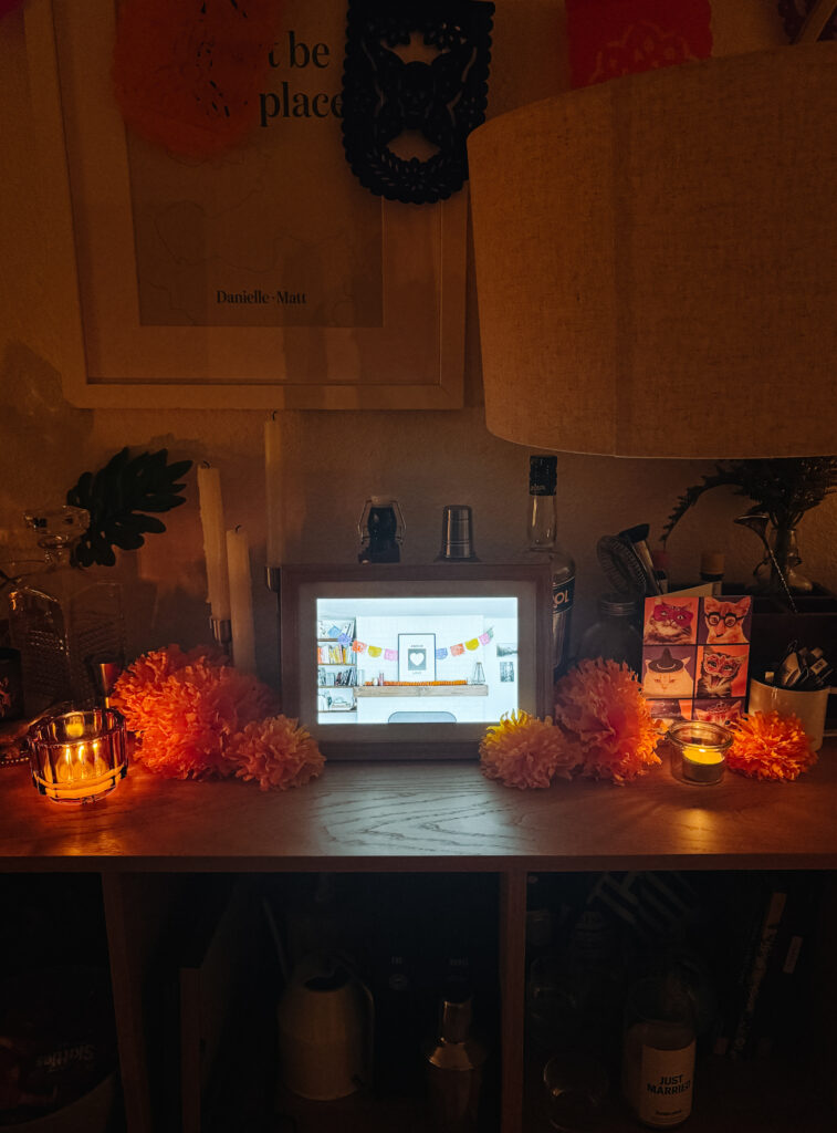 This image shows a cozy, dimly lit room with a wooden shelf holding various items. On the shelf, there is a framed digital display showing an interior scene, flanked by two lit candles and vibrant orange paper flowers. Above the shelf, a framed artwork with the text "be place" is mounted on the wall, accompanied by decorative elements. To the right, there is a lamp with a warm glow, casting a soft light over the scene. Below the shelf, several bottles and containers are stored, including one labeled "JUST MARRIED." The overall atmosphere is warm and inviting, with a focus on decorative and sentimental items.
