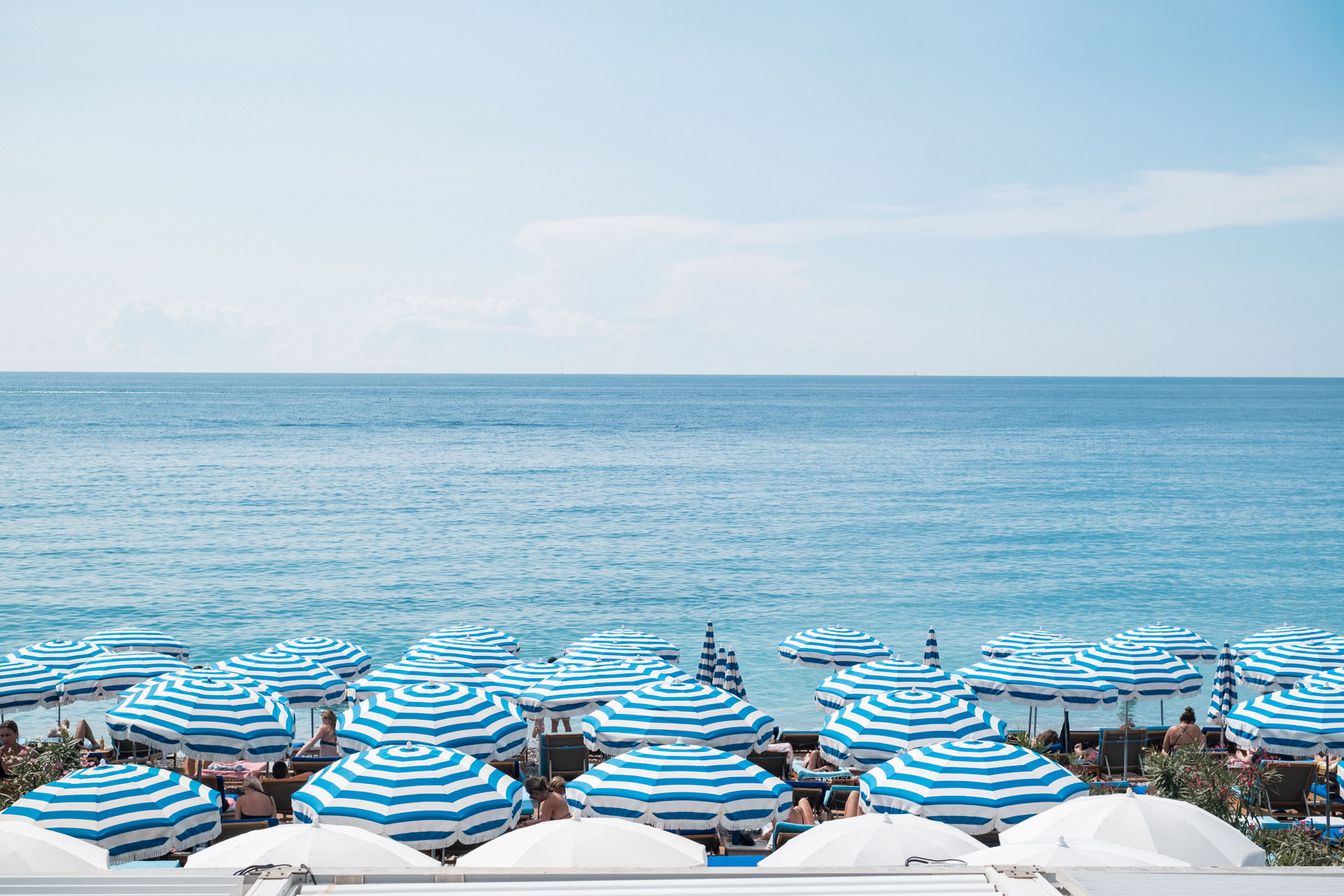 This image shows a beach scene with numerous blue and white striped umbrellas providing shade for people lounging on the sand. The beach is bordered by a clear blue ocean, with gentle waves visible. The sky above is mostly clear with a few scattered clouds. People are seen relaxing under the umbrellas, some lying on lounge chairs and others sitting. The overall atmosphere suggests a serene and enjoyable seaside environment.