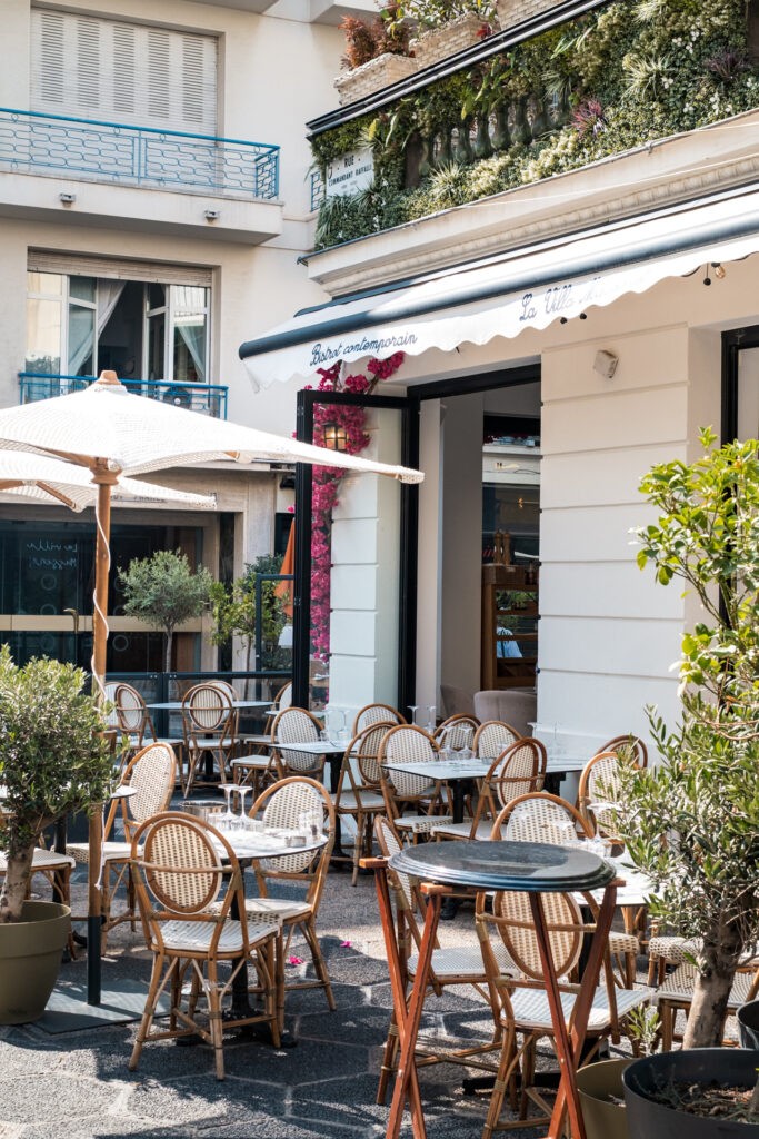 This image shows an outdoor dining area of a restaurant named "Bistrot contemporain" as indicated by the signage above the entrance. The restaurant features a modern design with a white awning and a lush green vertical garden on the facade. Several tables and chairs are arranged under large white umbrellas, providing shade. The chairs are made of wicker material, and the tables have wine glasses and other dining accessories on them. Potted plants are placed around the seating area, adding to the ambiance. The scene appears to be set in a vibrant urban environment, with the restaurant located on a street corner.