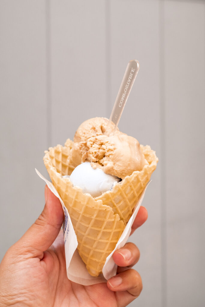This image shows a hand holding a waffle cone filled with two scoops of ice cream. The ice cream appears to be two different flavors, one light and one darker in color. A silver spoon is inserted into the cone, resting on top of the ice cream scoops. The cone is supported by a small piece of paper, which is held by the fingers of the hand. The background is plain and light-colored, providing a simple backdrop that highlights the ice cream and cone.