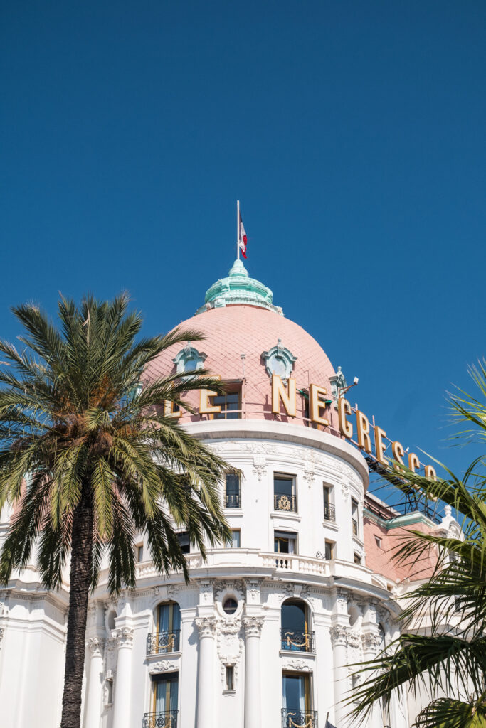 This image shows a prominent building with a distinctive pink dome and a flag at its peak. The building is adorned with ornate white facades and balconies, featuring decorative elements and railings. Large palm trees are visible in the foreground, adding to the scenic view. The structure has large, golden letters spelling out "LE NECRESCA" across its upper section, indicating the name of the building. The sky above is clear and blue, suggesting a sunny day.