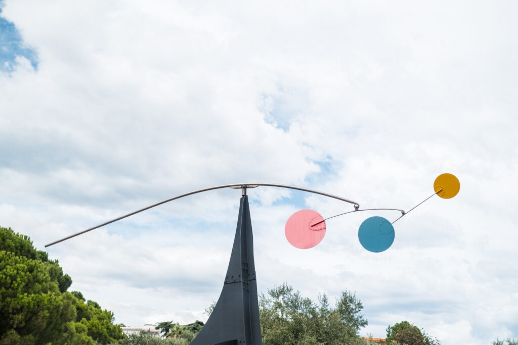 This image shows an abstract outdoor sculpture with a long, curved metal arm extending from a tall, triangular base. The arm holds three circular elements: one yellow at the far end, one red in the middle, and one blue closer to the base. The sculpture is set against a backdrop of a partly cloudy sky and is surrounded by green trees, suggesting it is located in a park or garden. The overall design appears modern and artistic, emphasizing balance and movement.