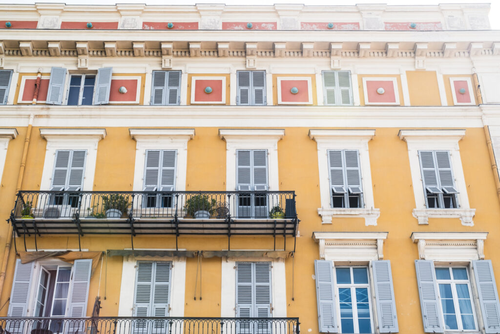 This image shows a multi-story building with a bright yellow facade and white architectural details. The building features numerous windows with white shutters, some of which are open, allowing a glimpse of the interiors. On the second floor, there is a balcony with a black metal railing, adorned with a few potted plants. The balcony is supported by decorative brackets. The building's design includes classical elements such as cornices and pediments, giving it a historic and elegant appearance.