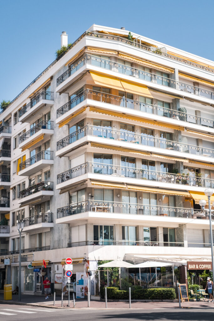 This image shows a multi-story building with numerous balconies and yellow awnings. The building has a modern architectural style with large glass windows. At the ground level, there are several storefronts, one of which has a sign reading "BAR & COFFEE." There are people walking on the sidewalk in front of the building, and some are seated at outdoor tables under umbrellas. The scene is set under a clear blue sky, suggesting a pleasant day.