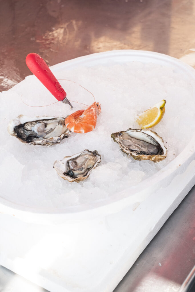This image shows a close-up view of a white tray filled with ice, on which several oysters and a shrimp are placed. The oysters are shucked, revealing their meat inside the shells. A red oyster knife with a textured handle is positioned on the ice, likely used for opening the oysters. A slice of lemon is also present on the ice, possibly for adding flavor to the seafood. The tray is placed on a metallic surface, which reflects some light.