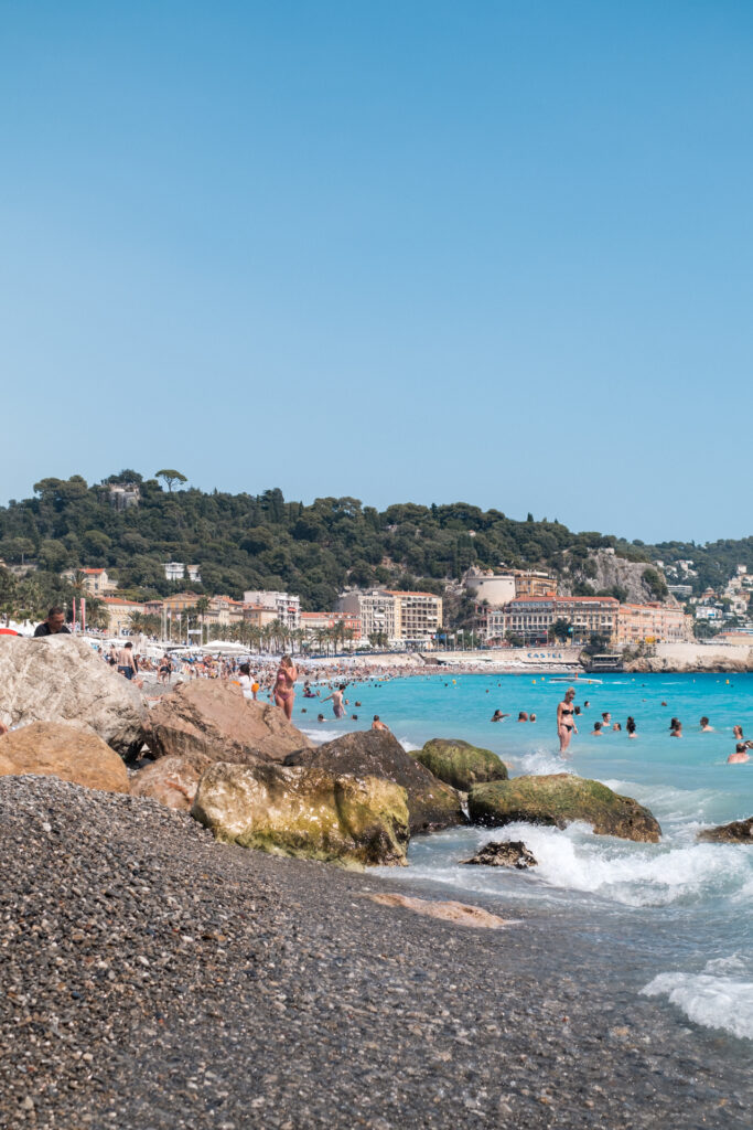 This image shows a bustling beach scene with numerous people enjoying the water and the shore. The beach is lined with large rocks and pebbles, and the water is a vibrant blue with small waves. In the background, there is a hill covered with trees and several buildings, some of which have the word "CASTEL" visible on them. The sky is clear and blue, indicating a sunny day. People are seen swimming, standing, and walking along the beach, creating a lively atmosphere.