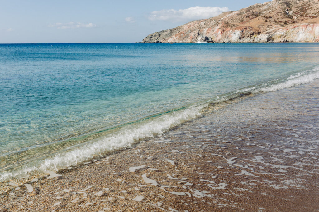 This image shows a serene beach scene with clear blue waters gently lapping onto the shore. The beach has a mix of sand and small pebbles, with the water creating a white foamy edge as it reaches the land. In the background, there is a rugged, rocky coastline with varied terrain, including cliffs and slopes. The sky above is mostly clear with a few scattered clouds. The overall atmosphere suggests a tranquil and picturesque coastal environment.