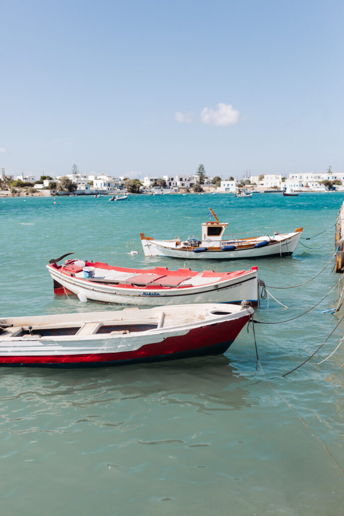This image shows three small boats floating on a calm body of water, likely a sea or a large lake. The boats are moored close to each other with ropes. The boat in the foreground is predominantly white with red accents and has the text "Nikita" visible on its side. The middle boat is also white with red accents and displays the text "MAYPOMATA A.M.1368". The third boat, slightly further back, is white with blue accents. In the background, there is a coastal town with numerous white buildings and some greenery, suggesting a Mediterranean or tropical location. The sky is clear with a few scattered clouds.
