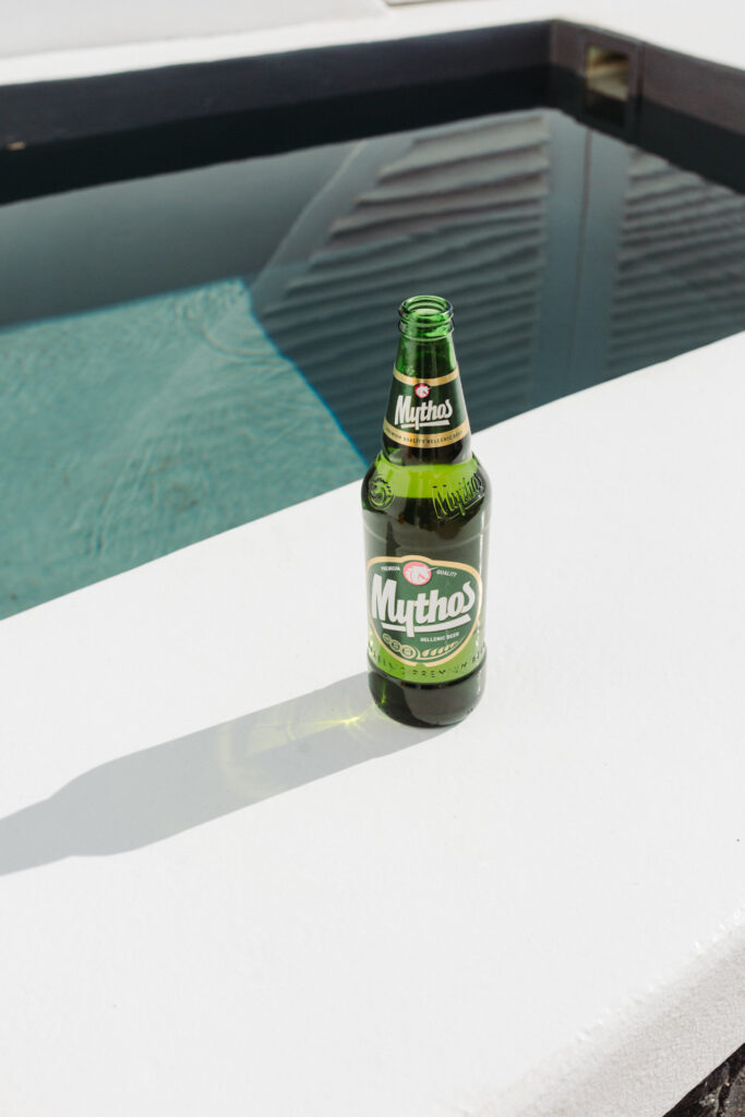 This image shows a bottle of Mythos beer placed on the edge of a swimming pool. The bottle is green and has the Mythos logo prominently displayed on the label. The label also includes the text "PREMIUM QUALITY HELLENIC BEER" and other descriptive phrases. The swimming pool water is visible in the background, reflecting light and creating a serene atmosphere. The bottle casts a shadow on the pool's edge, indicating bright sunlight.