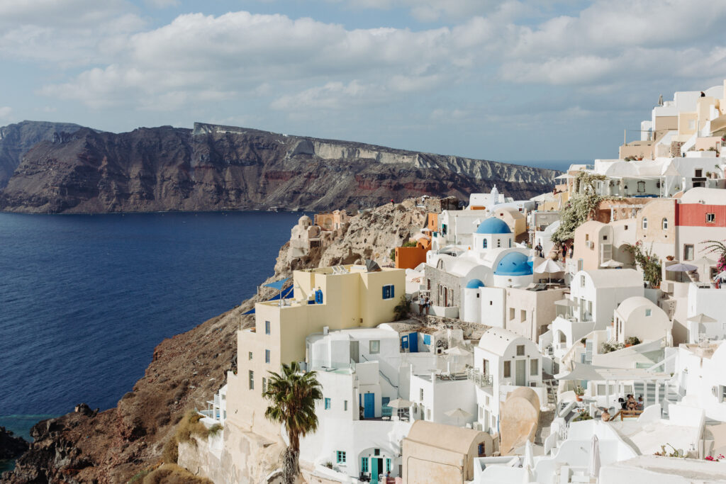 This image shows a picturesque coastal village with whitewashed buildings and blue-domed roofs, situated on a cliff overlooking the deep blue sea. The village is characterized by its traditional Cycladic architecture, with structures built close together and terraced on the hillside. The buildings are primarily white, with some featuring orange and yellow accents. The scene is set against a backdrop of a large, rugged cliff and a partly cloudy sky. Palm trees and other vegetation add a touch of greenery to the landscape.
