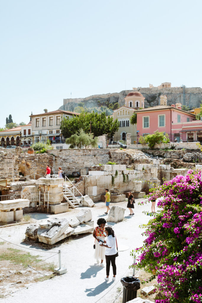 This image shows an outdoor scene with several people exploring an ancient site. The site features stone ruins and remnants of old structures, suggesting a historical or archaeological location. In the background, there are modern buildings, including a pink house with a distinctive architectural style. A group of people are walking around the ruins, some are standing near a large bush with purple flowers on the right side of the image. The sky is clear and bright, indicating a sunny day.