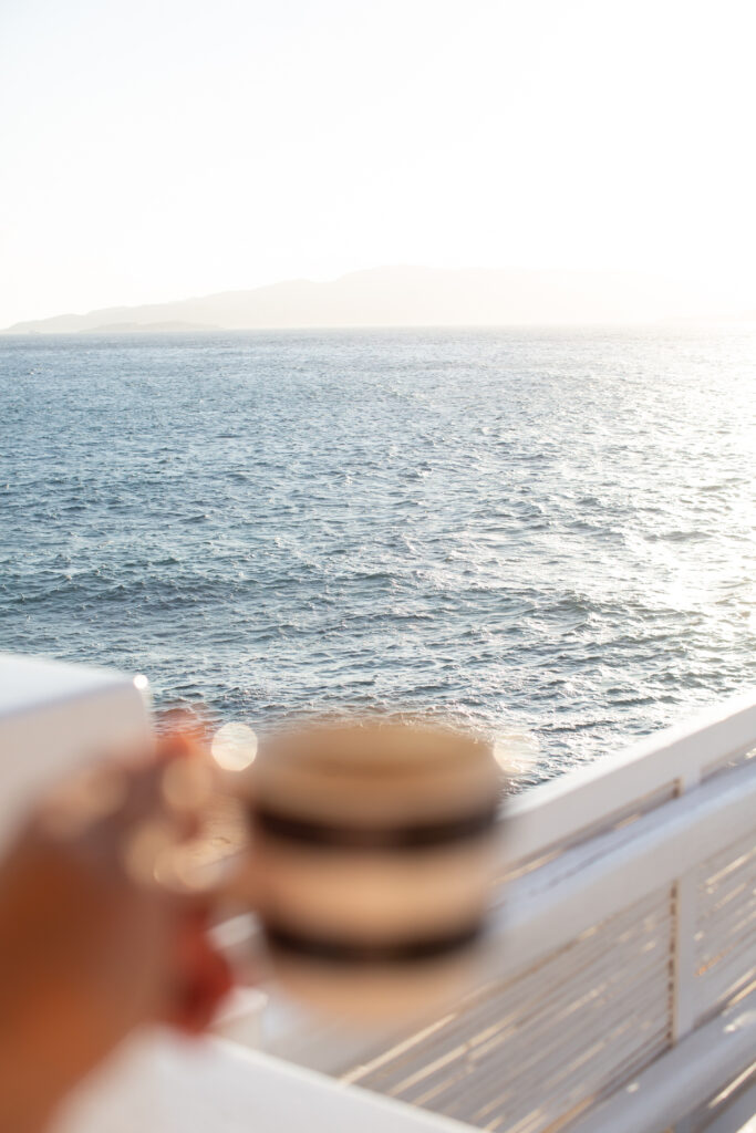 This image shows a serene scene where a person is holding a cup with a striped pattern, possibly enjoying a beverage. The cup is held close to the camera, creating a blurred effect. The background reveals a balcony with a white railing, overlooking a vast, calm ocean. The ocean stretches out under a clear sky, with a distant silhouette of mountains on the horizon. The overall atmosphere suggests a moment of relaxation and tranquility by the sea.