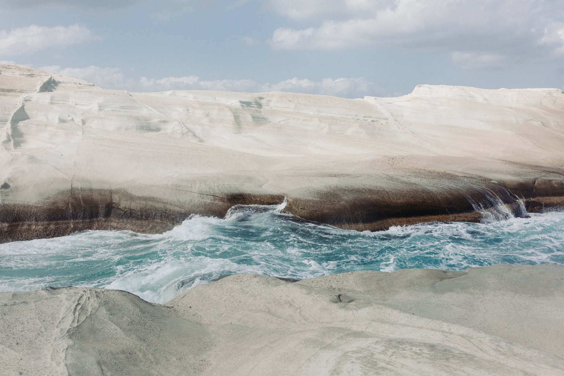 This image shows a dramatic coastal landscape with rugged, white cliffs towering above the ocean. The cliffs are composed of layered rock formations, some of which appear to be crumbling into the sea. The ocean below is turbulent, with waves crashing against the cliffs, creating a dynamic and powerful scene. The sky above is partly cloudy, with patches of blue visible, suggesting fair weather. The overall atmosphere is one of natural beauty and raw power, highlighting the forces of erosion and the relentless movement of the sea.