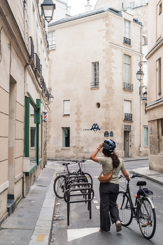 This image shows a narrow street with buildings on both sides. A person is walking on the street, wearing a helmet and carrying a brown bag. Several bicycles are parked on the sidewalk, secured to a bike rack. The buildings have multiple windows and balconies, with some having lamps mounted on the walls. There is a barber shop sign on the left side of the street. The street appears to be in a residential or mixed-use urban area.