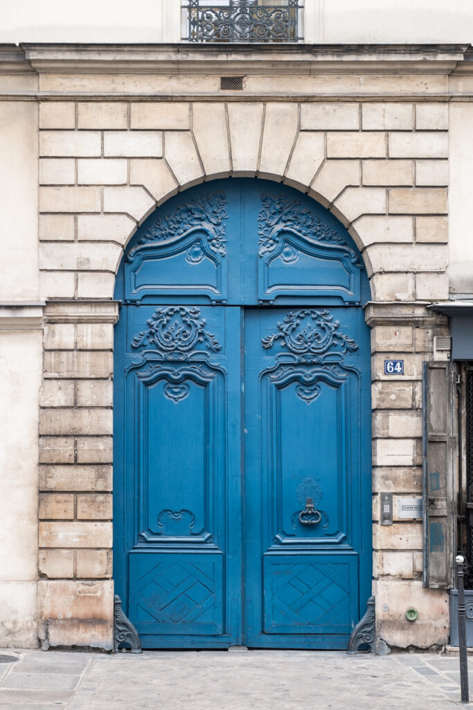 This image shows a large, ornate blue double door set within an arched entrance of a building. The door features intricate designs and patterns, including floral motifs and geometric shapes. A black door knocker is positioned near the bottom center of the door. Above the door, there is a balcony with a decorative metal railing. To the right of the door, a small sign with the number "64" is visible, indicating the building's address. The building's facade is constructed with light-colored stone bricks.