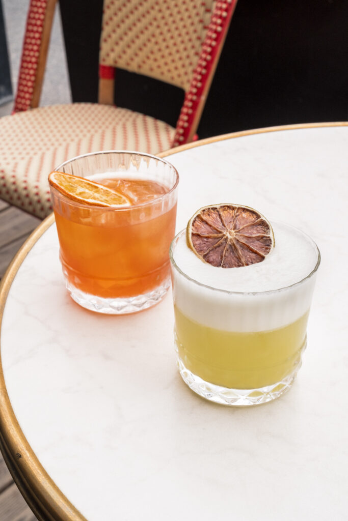 This image shows two glasses of alcoholic beverages placed on a round table with a marble top. The glass on the left contains an amber-colored drink topped with a slice of orange. The glass on the right contains a lighter-colored drink with a frothy head and a slice of dried orange on top. The table is set outdoors, with a bistro chair visible in the background, featuring a red and white striped design. The scene suggests a casual, relaxed setting, possibly at a bar or a cafe.