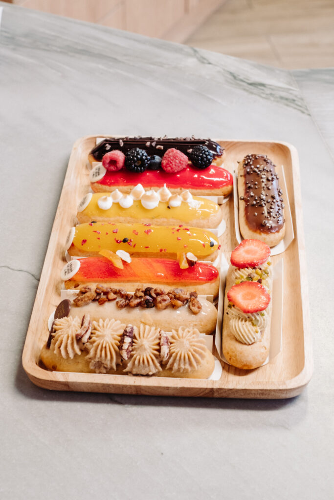 This image shows a wooden tray filled with an assortment of pastries. The pastries include éclairs with various toppings such as berries, whipped cream, nuts, and chocolate. Each pastry is labeled with the text "L'ÉCLAIR" and "DE GENE" indicating the type and possibly the brand or origin. The tray is placed on a light-colored surface, likely a table or countertop. The scene suggests a presentation of gourmet desserts, possibly in a bakery or café setting.