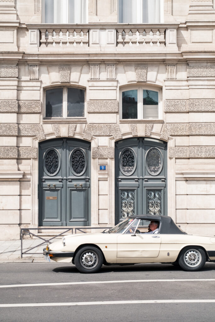 This image shows a classic car parked in front of an ornate building. The car is a convertible with a beige exterior and a black top. A person is seated in the driver's seat, wearing a white shirt. The building features intricate architectural details, including decorative stonework and two large, ornate doors. Above the doors, there are two windows with decorative frames. A small blue sign with the number "2" is visible near the doors. The scene appears to be set in an urban environment with a paved road in front of the building.