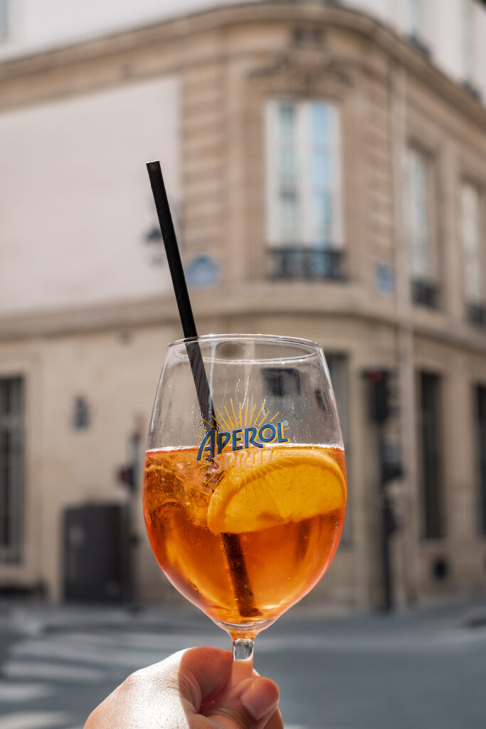 This image shows a hand holding a wine glass filled with a beverage that appears to be an Aperol Spritz, indicated by the Aperol logo visible on the glass. The drink is a light orange color with a slice of lemon floating inside. A black straw is inserted into the drink. The background features a street scene with a building that has multiple windows and balconies. The setting suggests an outdoor environment, possibly in a city.