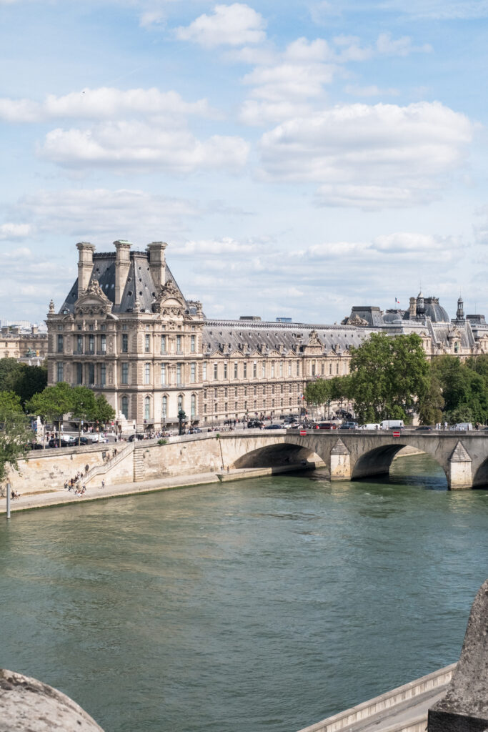 This image shows a picturesque view of the Seine River in Paris, with the historic Louvre Palace prominently visible in the background. The river is calm and stretches across the foreground, with a stone bridge crossing it. The bridge is bustling with pedestrians and vehicles, indicating it is a major thoroughfare. The Louvre Palace, with its grand architecture and numerous windows, stands majestically along the riverbank. Trees and streetlights line the pathways on both sides of the river, adding to the scenic beauty. The sky above is partly cloudy, with patches of blue visible.