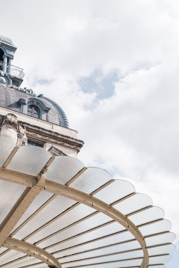 This image shows an architectural scene featuring a building with a dome-shaped roof and intricate detailing. The building has a classical design with ornate elements such as decorative moldings and a balcony with a wrought-iron railing. The sky above is partly cloudy, with patches of blue visible. In the foreground, there is a transparent, curved canopy structure with a metallic frame, possibly part of an entrance or a decorative feature. The overall scene suggests a historical or grand architectural style.