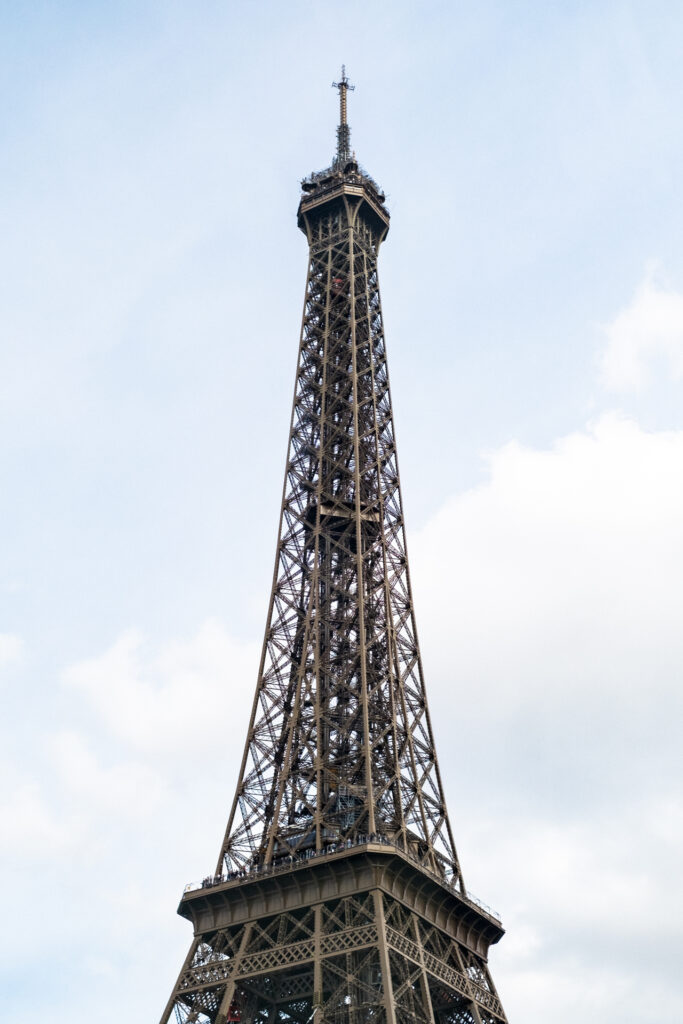 This image shows the Eiffel Tower, a wrought iron lattice tower located in Paris, France. The tower is tall and slender, tapering as it rises, with a distinct lattice structure that is characteristic of its design. The sky in the background is mostly clear with some scattered clouds. The tower appears to have a few people on its upper levels, enjoying the view from the observation decks. The overall scene is iconic and represents a famous landmark known worldwide.
